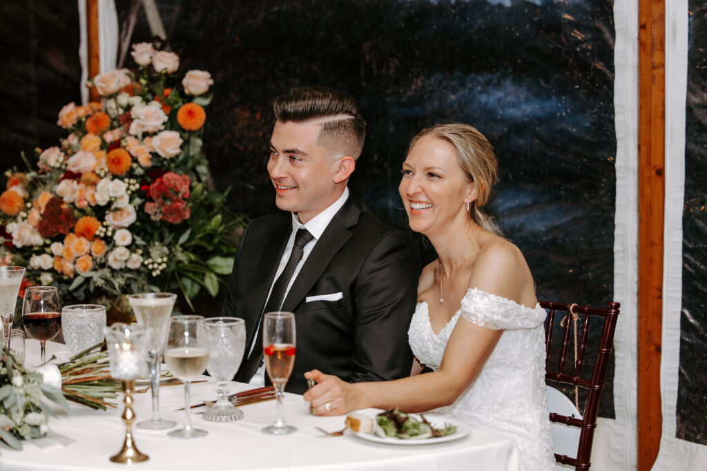 Bride and groom smile while their wedding party gives speeches during their fall wedding at The Willodale Estate.