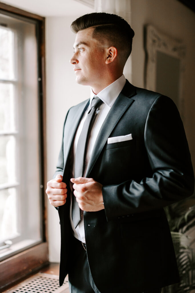 Groom looks out the window while holding suit jacket after getting ready for his wedding at The Willowdale Estate.
