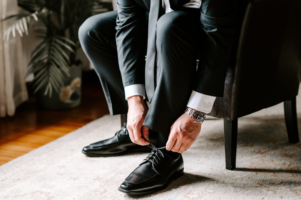Groom wearing a black suit ties his shoes while he gets ready for his wedding at The Willowdale Estate.