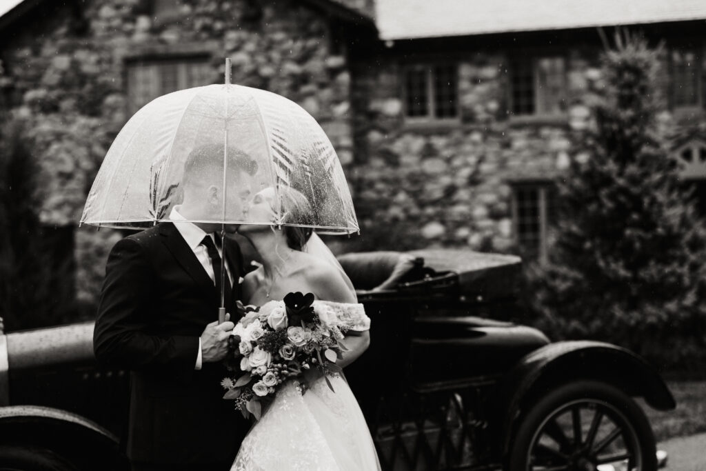 Bride and groom kiss under an umbrella on their rainy wedding day in front of The Willowdale Estate.