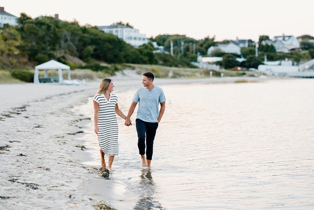 newly engaged couple hold hands and smile as they walk on the beach in Cape Cod