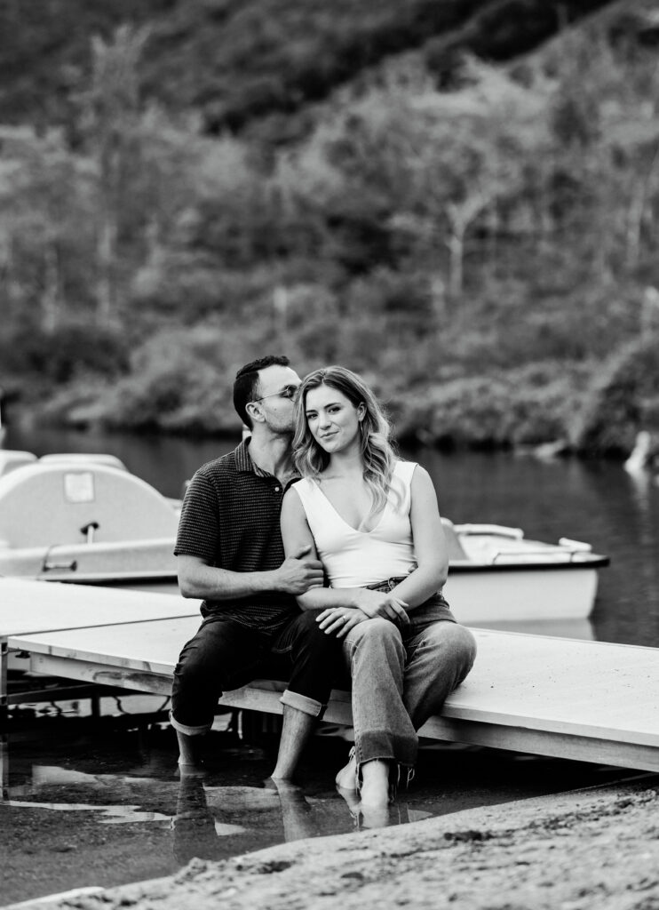 Groom kisses bride on boat dock towards NH wedding photographer during summer mountain engagement photos in New Hampshire
