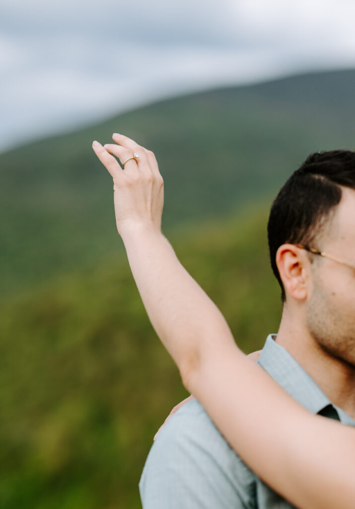 Bride holds engagement ring in the air in NH white mountains during summer mountain engagement photos in New Hampshire
