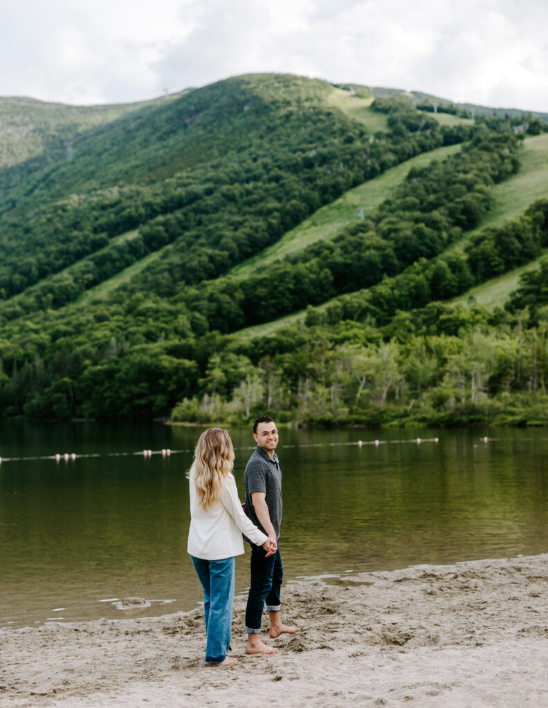 Groom smiles at camera and holds brides hand in front of Cannon Mountain in New Hampshire
