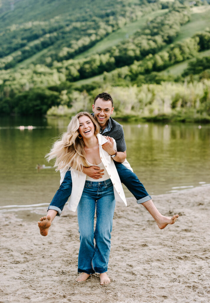 Bride holds groom on his back as they laugh towards NH wedding photographer during summer mountain engagement photos in New Hampshire