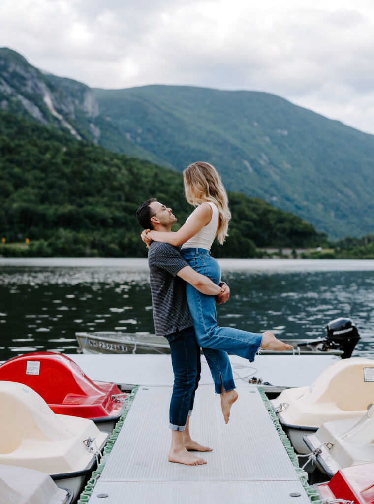 Man holds woman under the butt lift in NH white mountains during summer mountain engagement photos in New Hampshire
