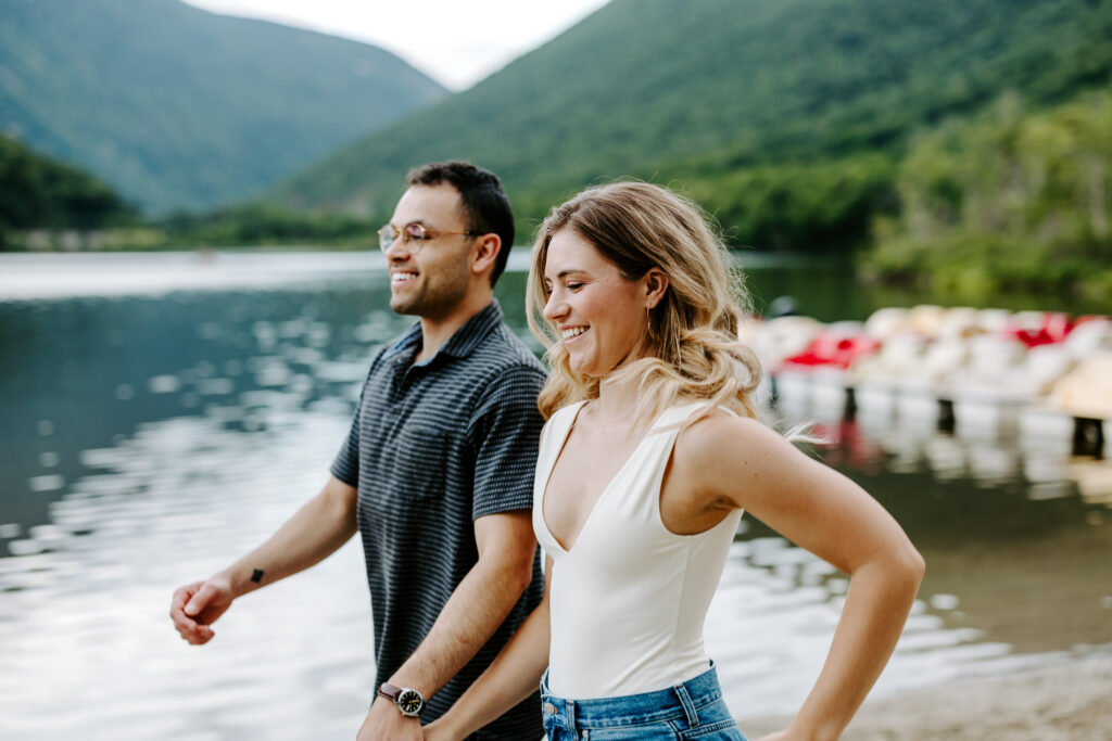 Couple holds hands and run down the beach during engagement photo session in Franconia Notch, Echo Lake beach.