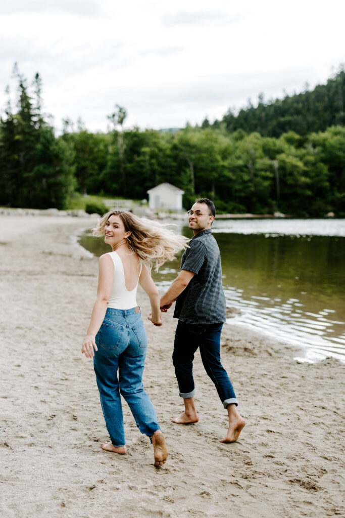 Bride smiles over shoulder while she runs down Echo Lake Beach with groom during summer mountain engagement photos in New Hampshire