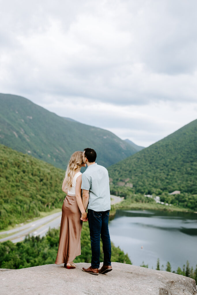 Groom kisses bride on head at the top of NH white mountains during summer mountain engagement photos in New Hampshire