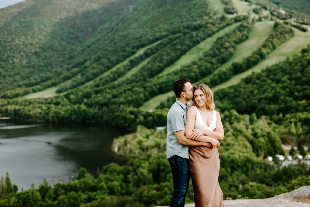 Groom wraps arms around and kisses bride at Echo Lake in NH during summer mountain engagement photos in New Hampshire