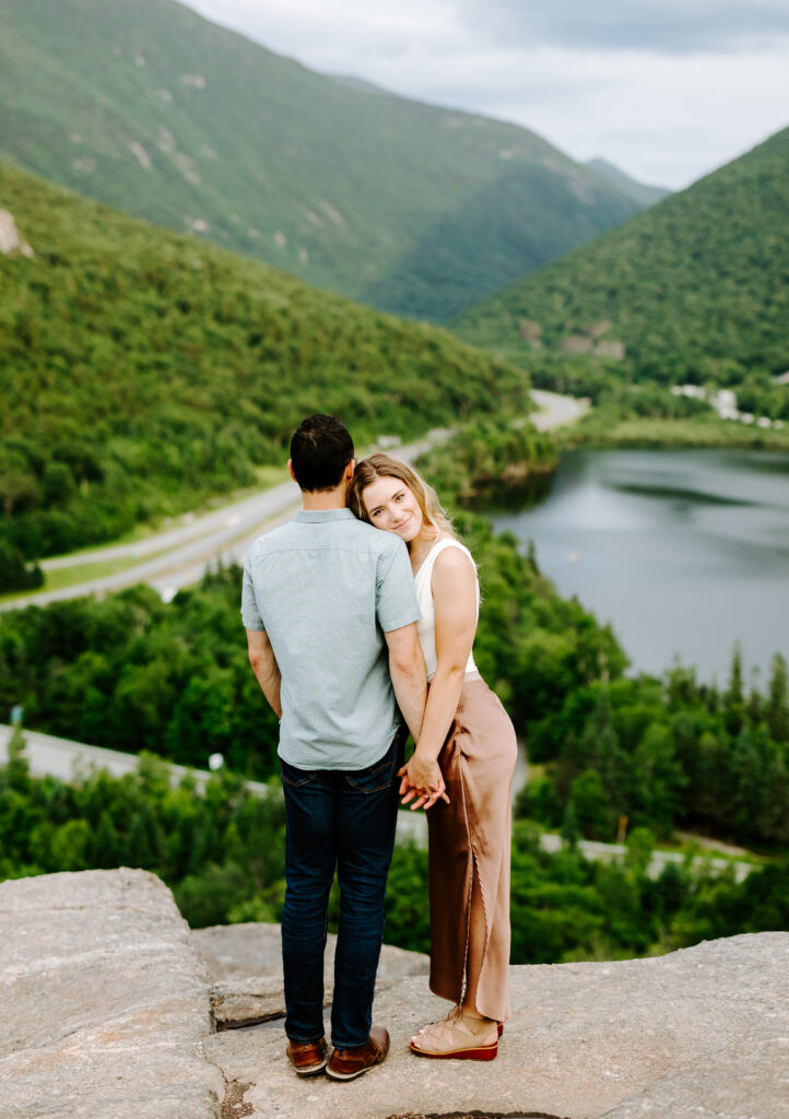 Bride smiles over grooms shoulder at NH wedding photographer in summer mountain engagement photos in New Hampshire
