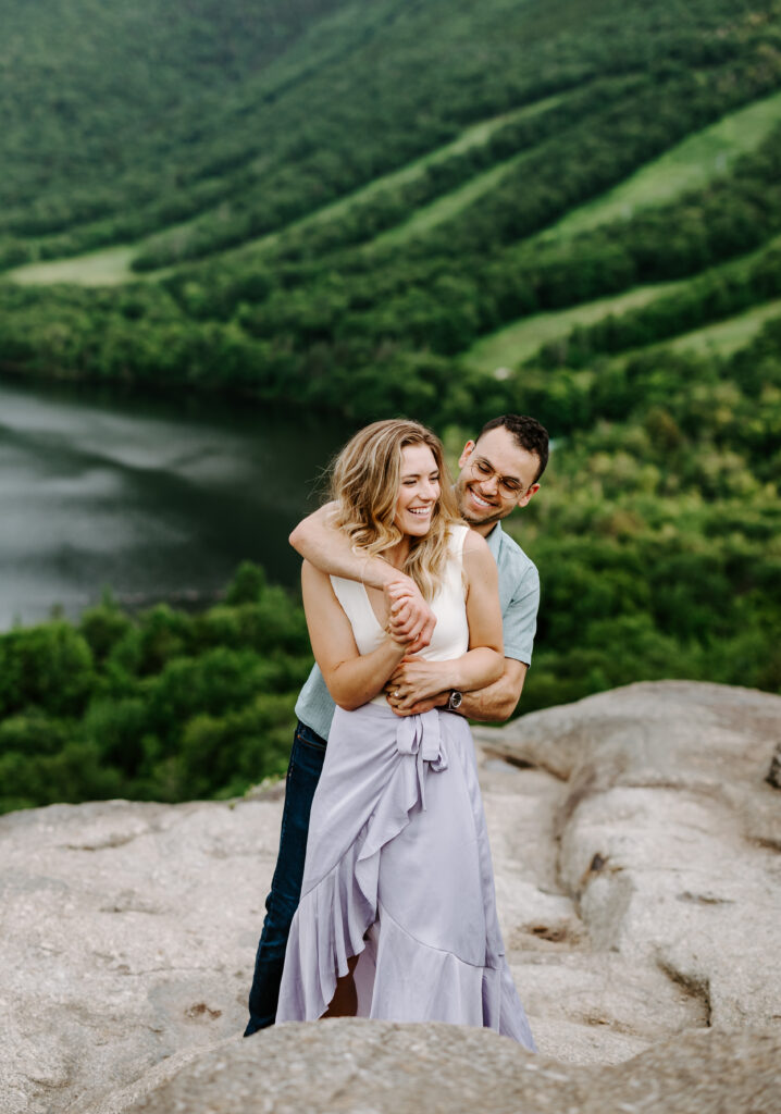 Bride and groom hold each other and laugh on top of mountain in NH during engagement session.
