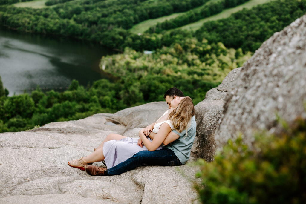 Bride and groom sit on top of mountain rock and hold each other in summer mountain engagement photos in New Hampshire
