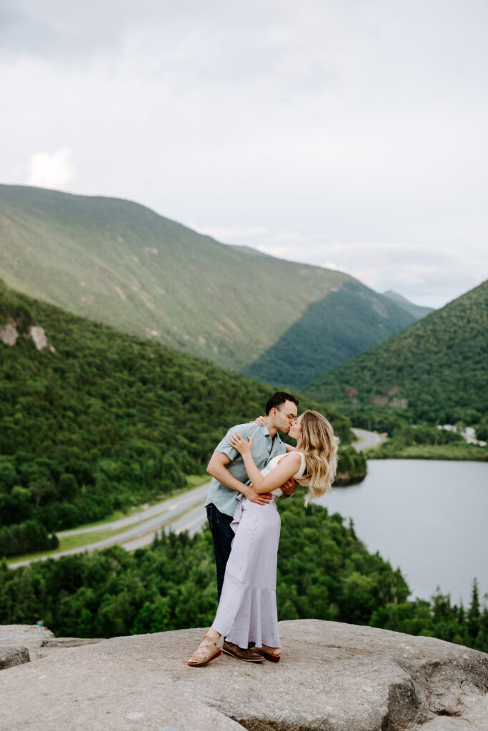 Bride and groom dip kiss on top of Artists Bluff in Franconia Notch in New Hampshire in summer mountain engagement photos in New Hampshire
