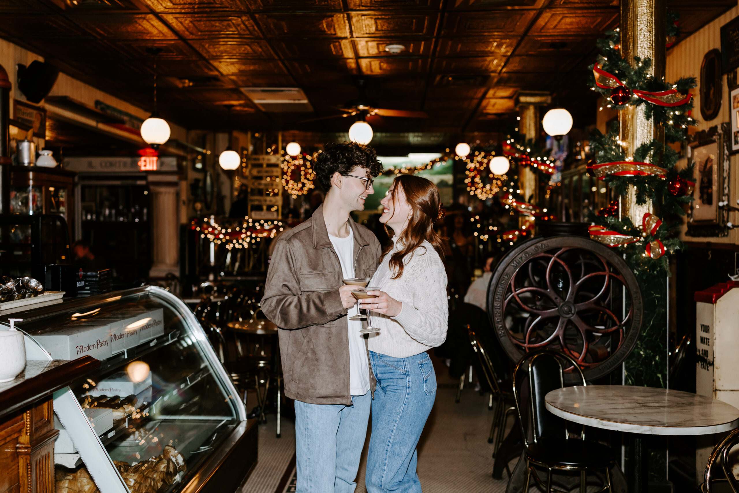 Couple smiles at each other with espresso martinis in cozy Boston cafe during engagement photo session