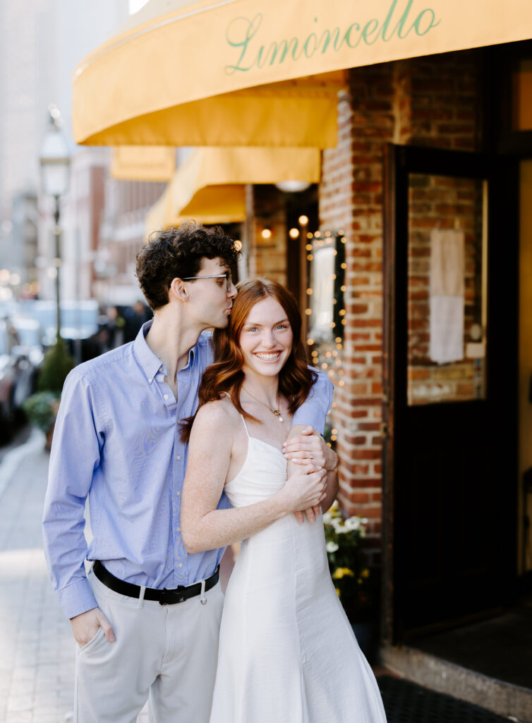 Groom kisses bride while she smiles during engagement session