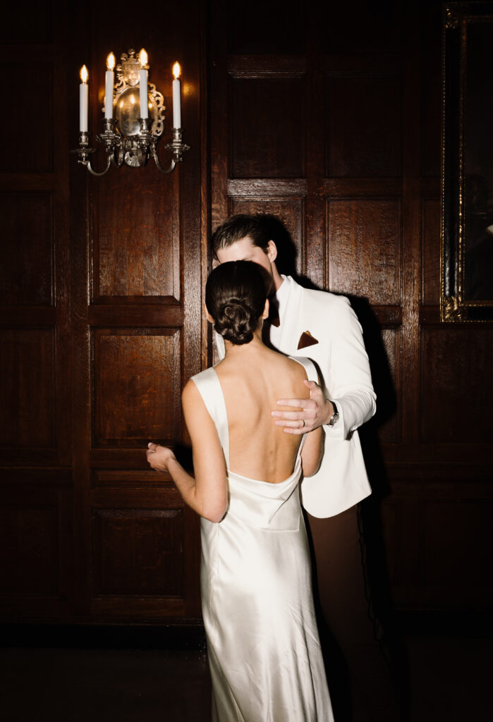 Groom holds and kisses bride after cutting cake at indoor wedding reception at The Boston Harvard Club in Massachusetts