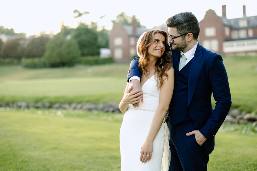 Grooms slings arm over bride's shoulder as they smile at each other in wedding photo in Massachusetts